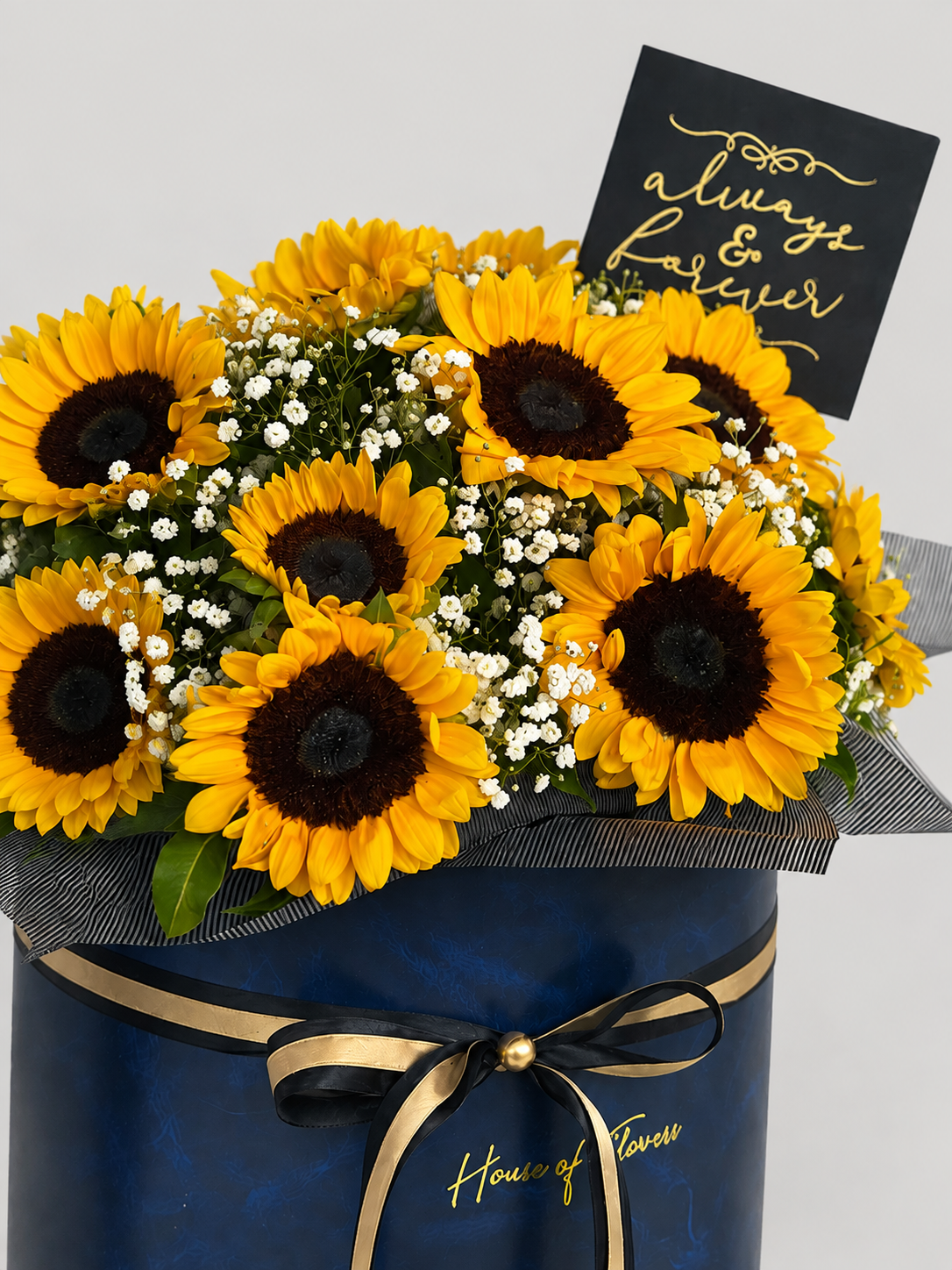 A vibrant bouquet of yellow sunflowers with dark brown centers and small white filler flowers, arranged in a dark blue cylindrical box with a gold ribbon and bow. A black card with gold cursive text reading &