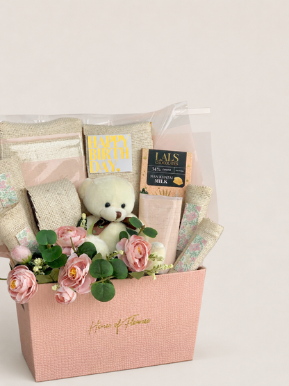 A pink gift basket arrangement from House of Flowers, featuring a small white teddy bear, pink roses, green leaves, burlap ribbon, a milk chocolate bar, and a card that reads &