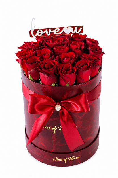 A round gift box filled with deep red roses, wrapped in a matching dark red ribbon with a pearl accent and gold script lettering. A small &