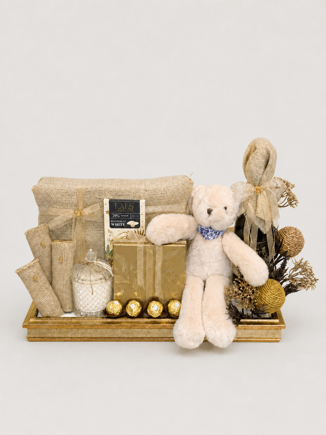 A styled gift basket arrangement on a gold tray, featuring a cream teddy bear with a blue bow, wrapped presents, dried botanicals, gold ornaments, and a small glass bottle, photographed against a light gray studio background.