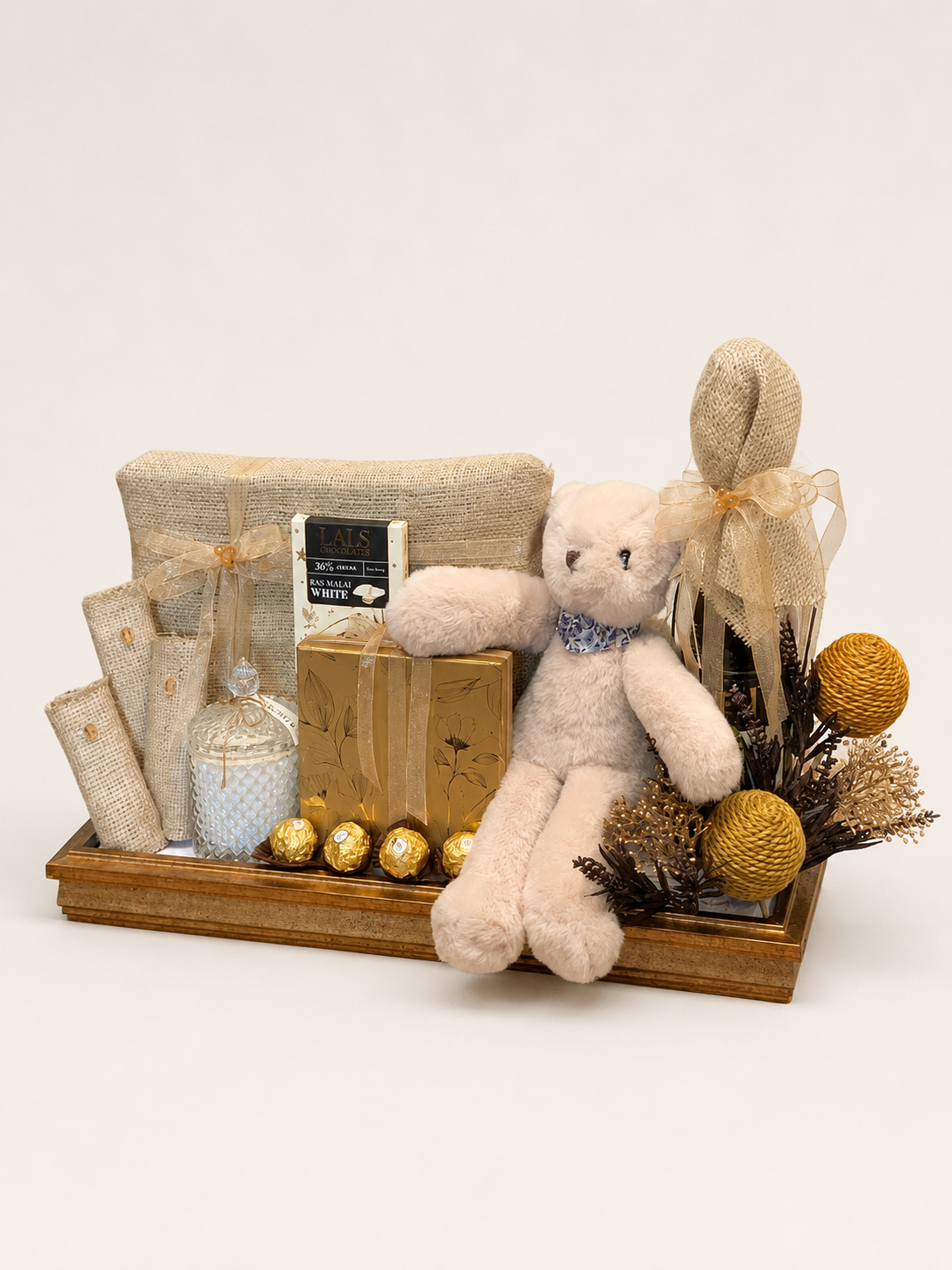 A rustic gift basket arrangement in a wooden tray, featuring a cream teddy bear with a blue bow, burlap-wrapped items, gold foil chocolates, and dried botanicals on a white background.