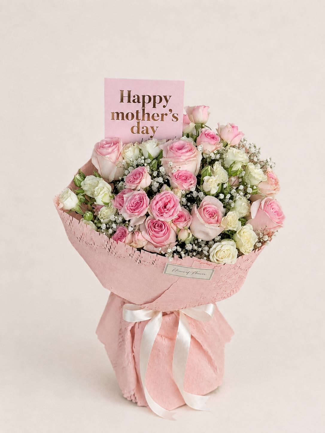 A pink and white rose bouquet wrapped in pink paper with a white ribbon, featuring a pink card that reads &