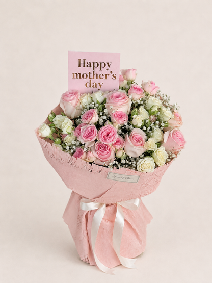 A pink and white rose bouquet wrapped in pink paper with a white ribbon, featuring a pink card that reads &