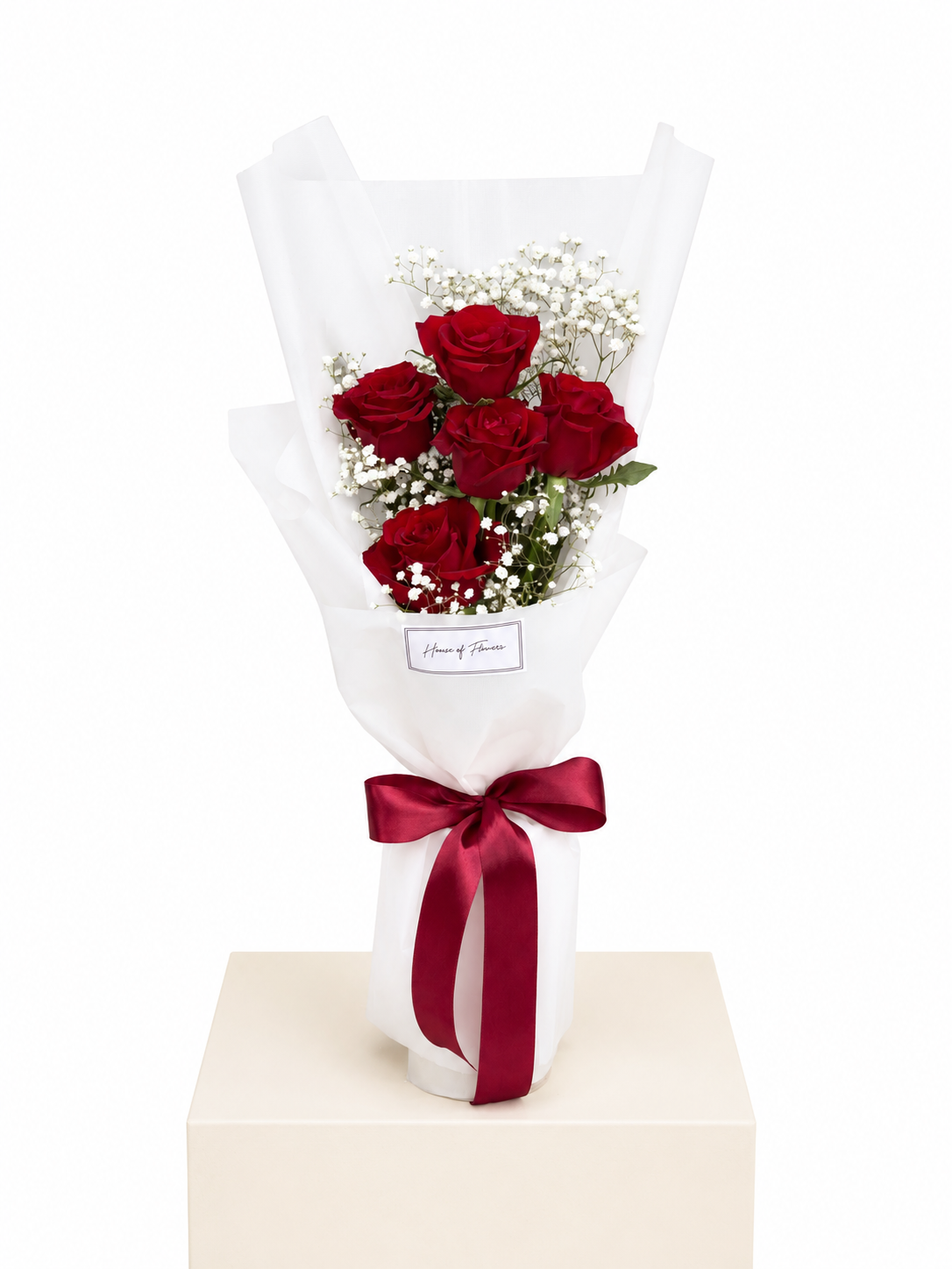 A bouquet of five red roses wrapped in white tissue paper with a dark red ribbon bow, displayed on a light beige pedestal against a white background.