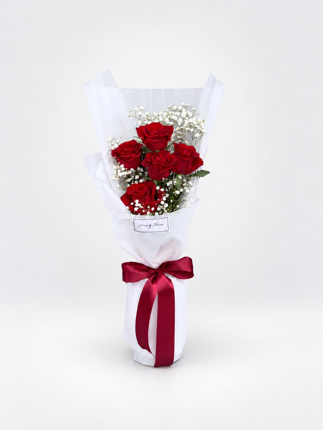 A bouquet of five red roses wrapped in white tissue paper with a dark red ribbon bow, displayed on a light beige pedestal against a white background.