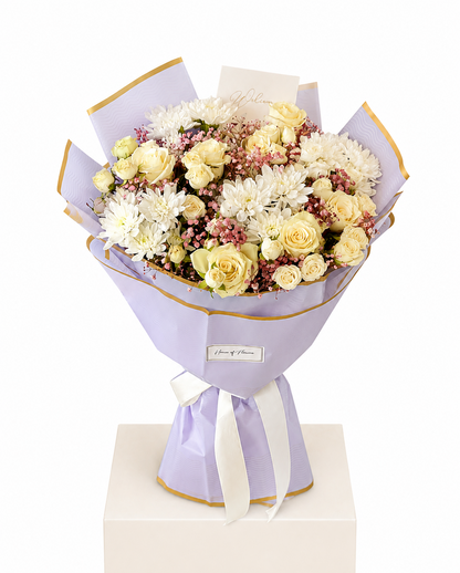 A floral bouquet wrapped in light lavender paper with gold trim and a white ribbon, displayed on a white pedestal against a white background. The bouquet contains cream and pale yellow roses, white chrysanthemums, and small pink filler flowers.