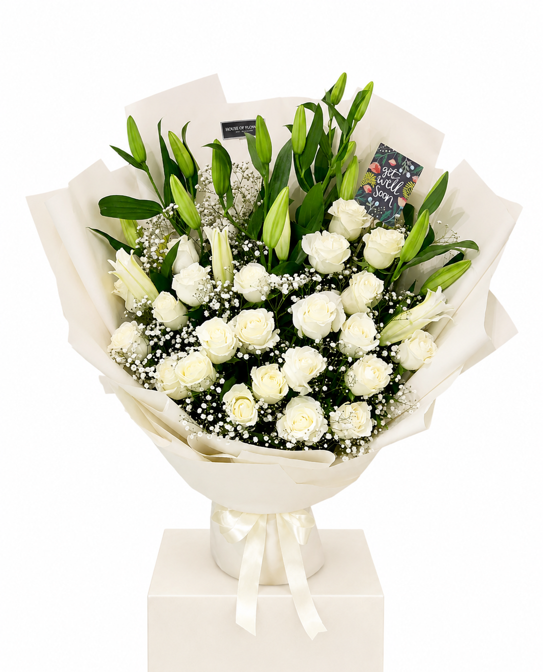 A bouquet of white roses and lilies with green leaves, wrapped in cream paper and tied with a white ribbon, displayed on a white box pedestal against a white background.