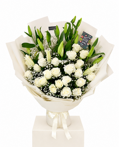 A bouquet of white roses and lilies with green leaves, wrapped in cream paper and tied with a white ribbon, displayed on a white box pedestal against a white background.