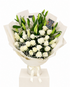 A bouquet of white roses and lilies with green leaves, wrapped in cream paper and tied with a white ribbon, displayed on a white box pedestal against a white background.