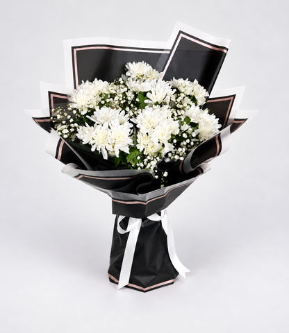A bouquet of white chrysanthemums wrapped in black paper with white ribbon accents, displayed on a light beige pedestal against a white background.