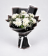 A bouquet of white chrysanthemums wrapped in black paper with white ribbon accents, displayed on a light beige pedestal against a white background.