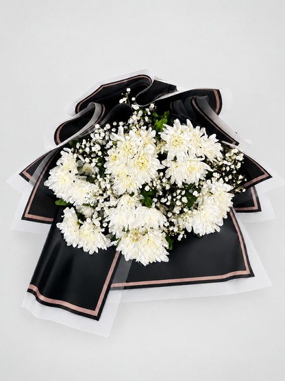 A bouquet of white chrysanthemums wrapped in black paper with white ribbon accents, displayed on a light beige pedestal against a white background.