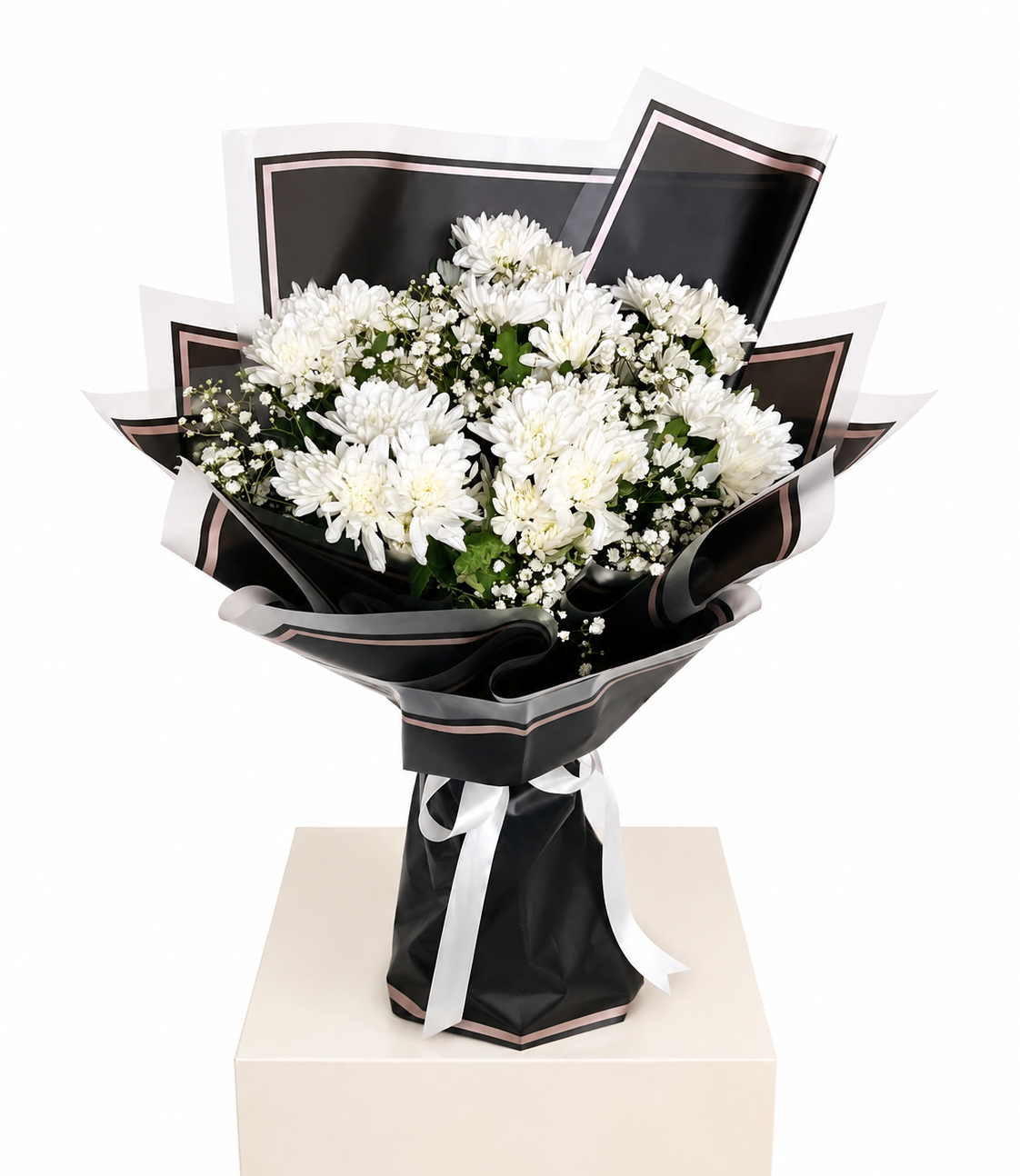 A bouquet of white chrysanthemums wrapped in black paper with white ribbon accents, displayed on a light beige pedestal against a white background.
