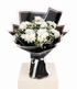 A bouquet of white chrysanthemums wrapped in black paper with white ribbon accents, displayed on a light beige pedestal against a white background.