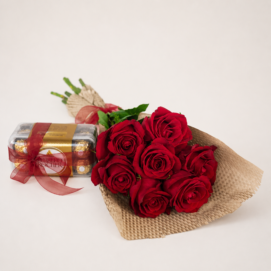 A bouquet of seven red roses wrapped in brown burlap with green stems and a red ribbon, placed next to a gold and red box of Ferrero Rocher chocolates on a plain off-white background.