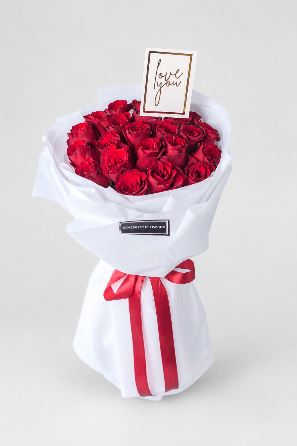 Bouquet of red roses wrapped in white paper with a red ribbon and a card on a white surface.