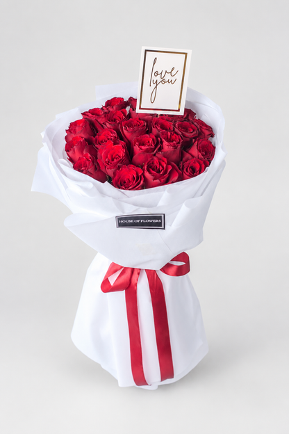 Bouquet of red roses wrapped in white paper with a red ribbon and a card on a white surface.