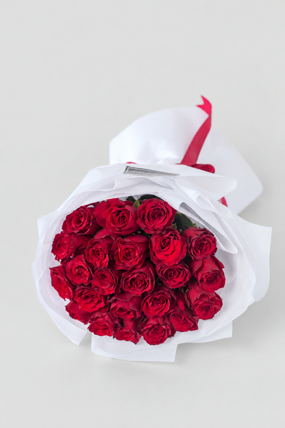 Bouquet of red roses wrapped in white paper with a red ribbon and a card on a white surface.